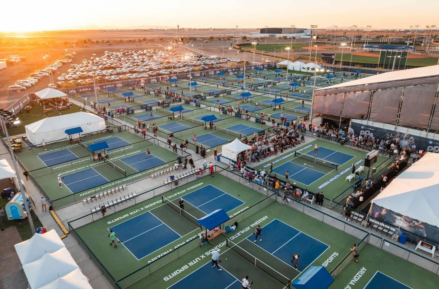 Aerial view of a large outdoor pickleball complex with many blue and green courts, players, and spectators under a sunset sky.