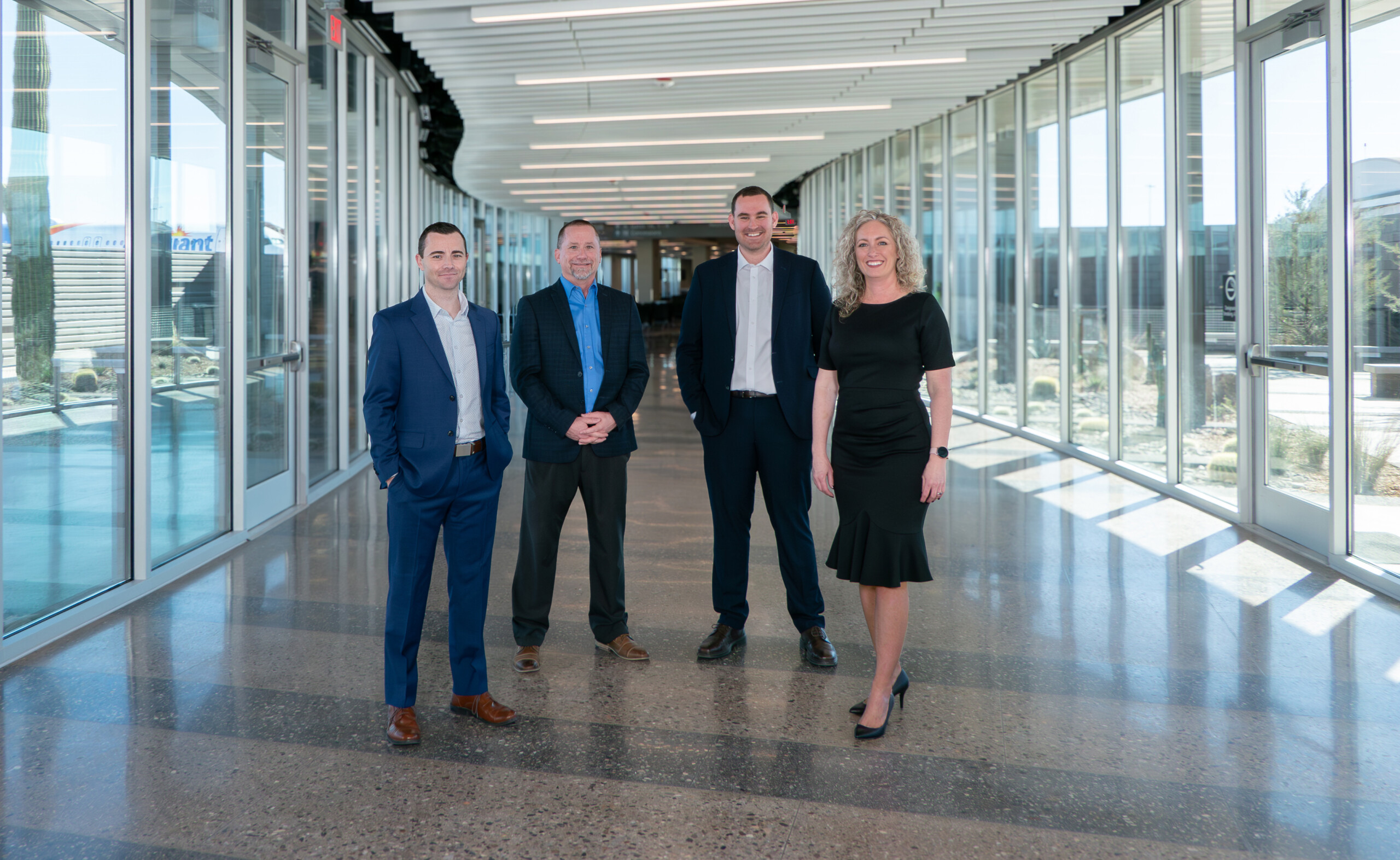 Four professionals, three men and one woman, stand smiling in a modern glass-walled hallway with a polished floor.