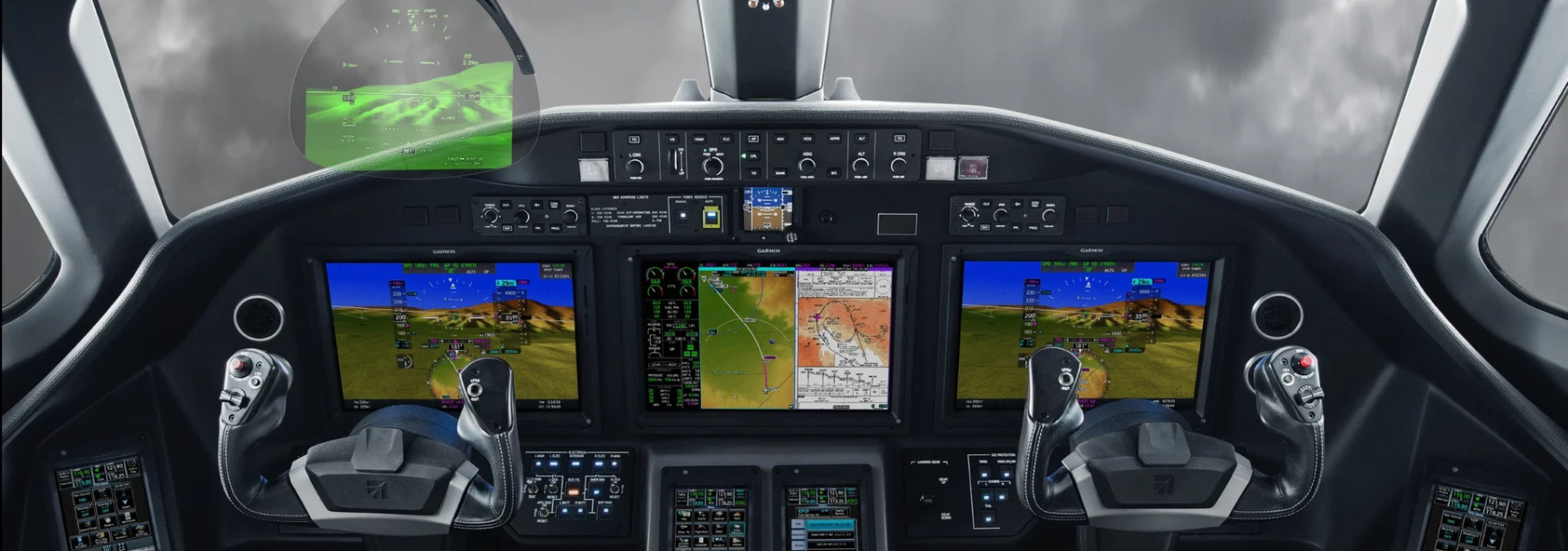 View from inside a modern aircraft cockpit showing three large digital displays, a head-up display, two side-stick control yokes, and numerous buttons and switches, with an overcast sky visible through the windshield.