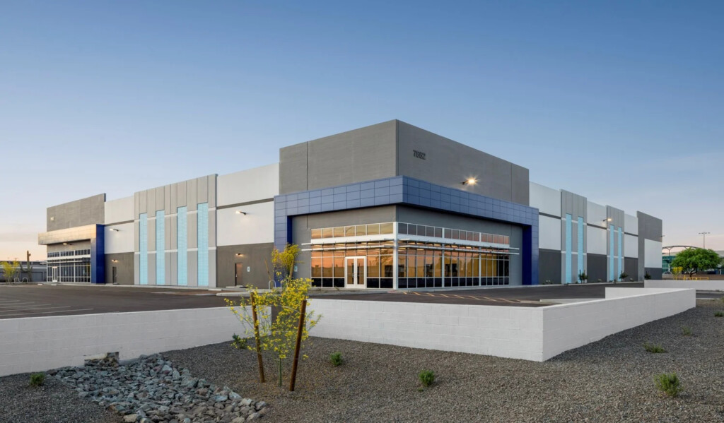 Exterior view of a large, modern commercial building with gray, white, and blue facade, glass entrance, and landscaped gravel foreground.