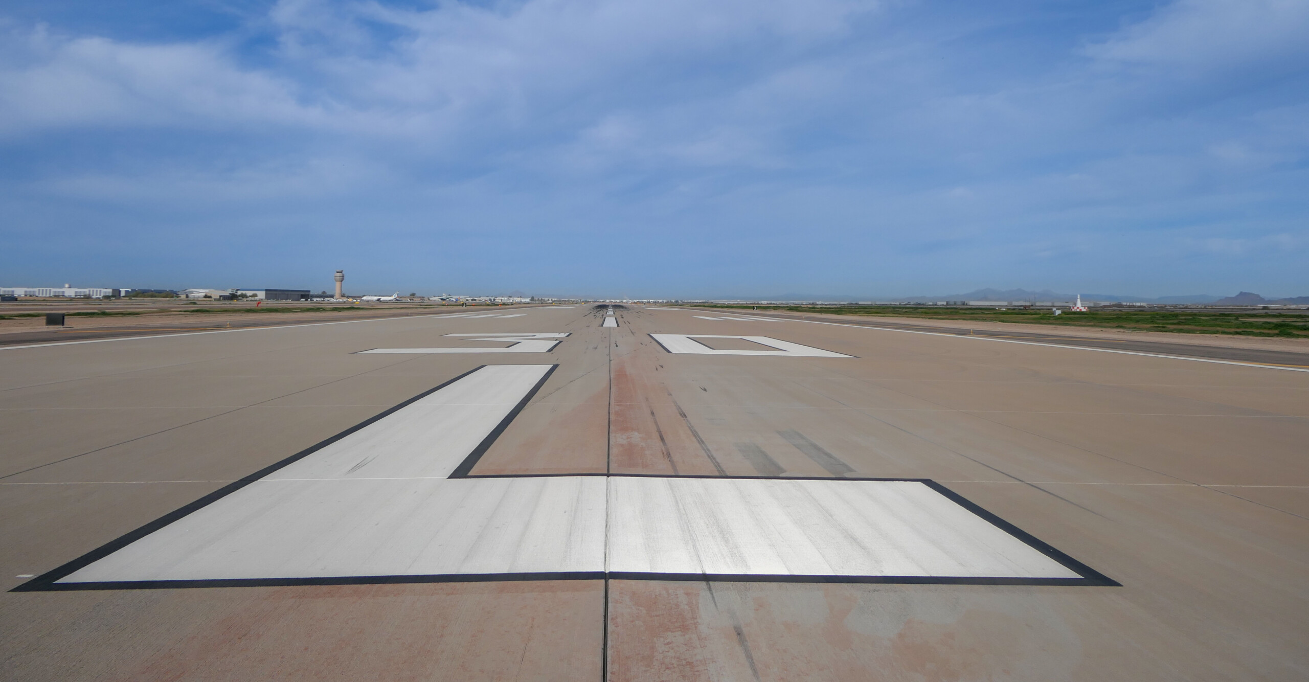An airport runway with white markings, a control tower, and mountains under a blue sky.