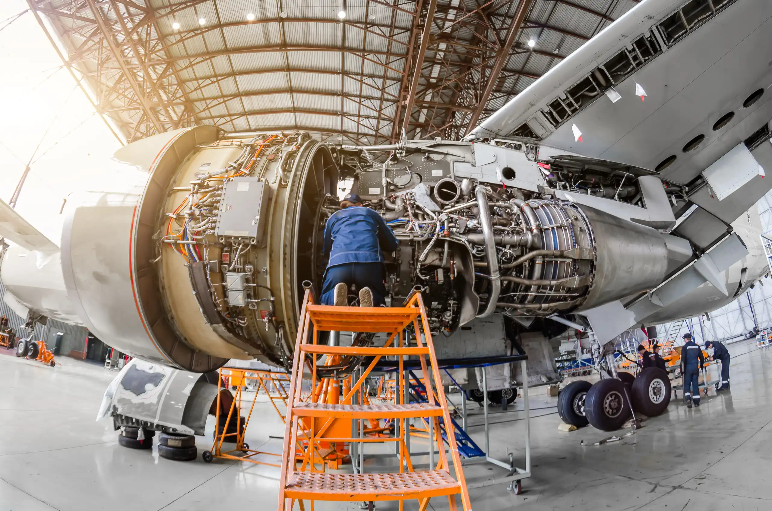 A mechanic in a blue uniform kneels on an orange platform, working on the exposed internal components of a large jet engine inside an aircraft hangar.