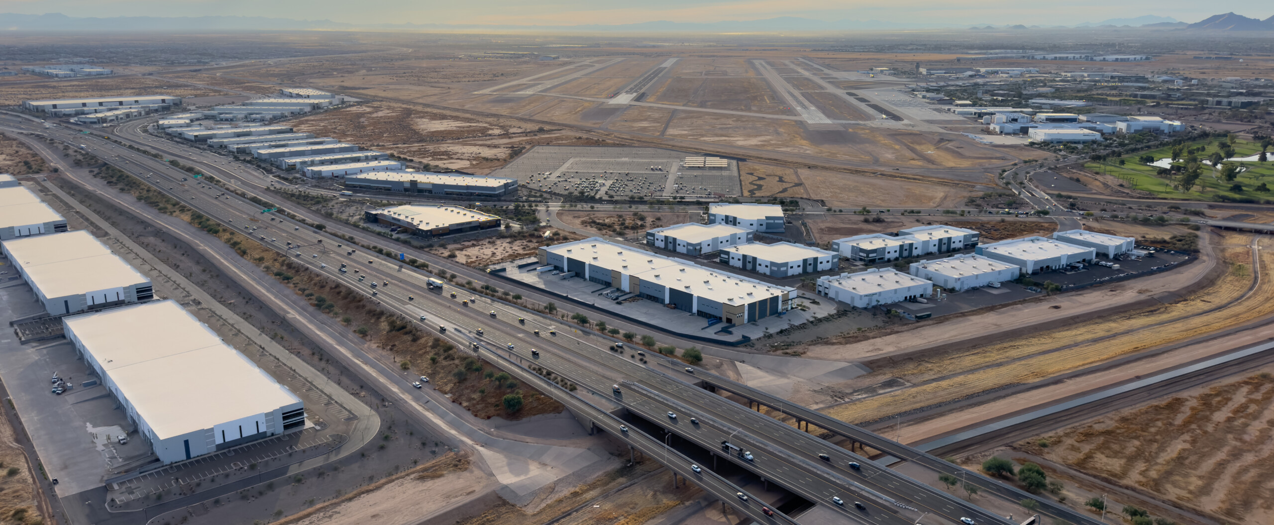 Aerial view of an industrial park with many large warehouses, a multi-lane highway with traffic, and an airport with runways, all set in a desert landscape under a hazy sky.