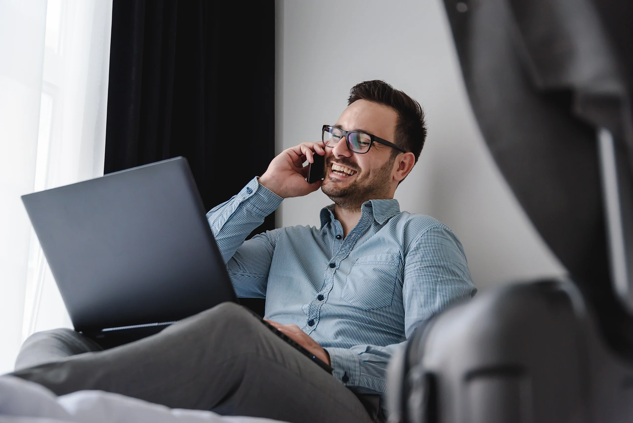A smiling man in glasses talks on a phone while using a laptop.