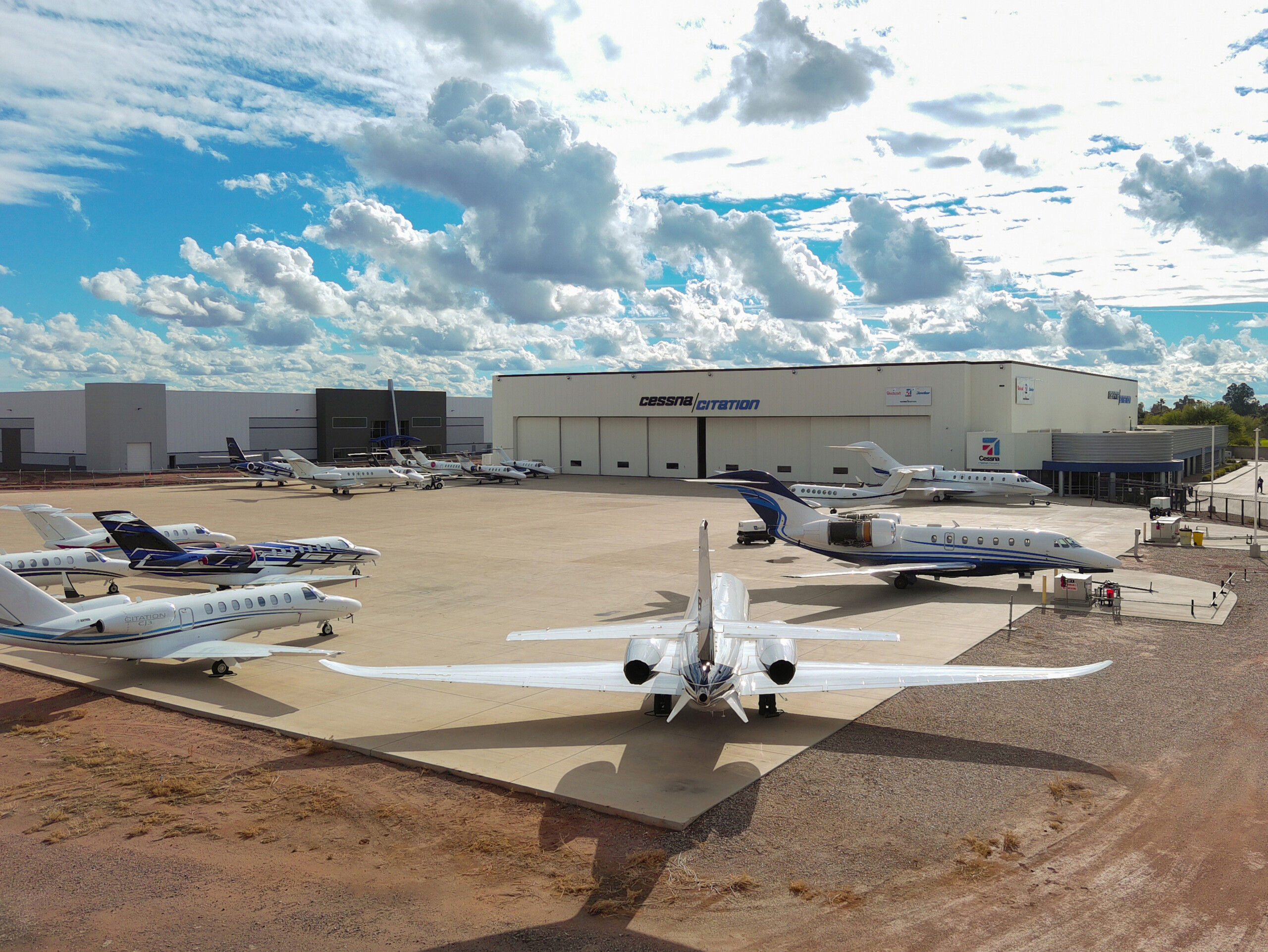 Aerial view of multiple white private jets parked on an airport apron in front of a large Cessna Citation hangar under a cloudy blue sky.