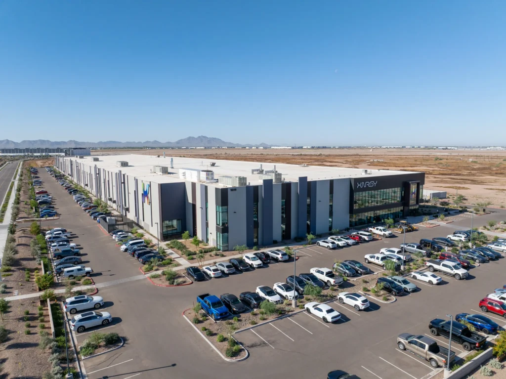Aerial view of a large, modern gray and black industrial building with 'XNERGY' logo, surrounded by parking lots full of cars, arid landscaping, and distant mountains under a clear blue sky.
