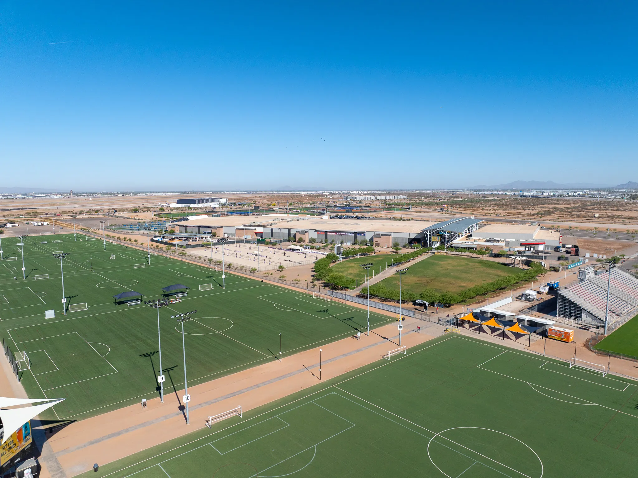 Aerial view of a large sports complex with multiple green soccer fields, stadium seating, and buildings under a clear blue sky in a desert landscape.