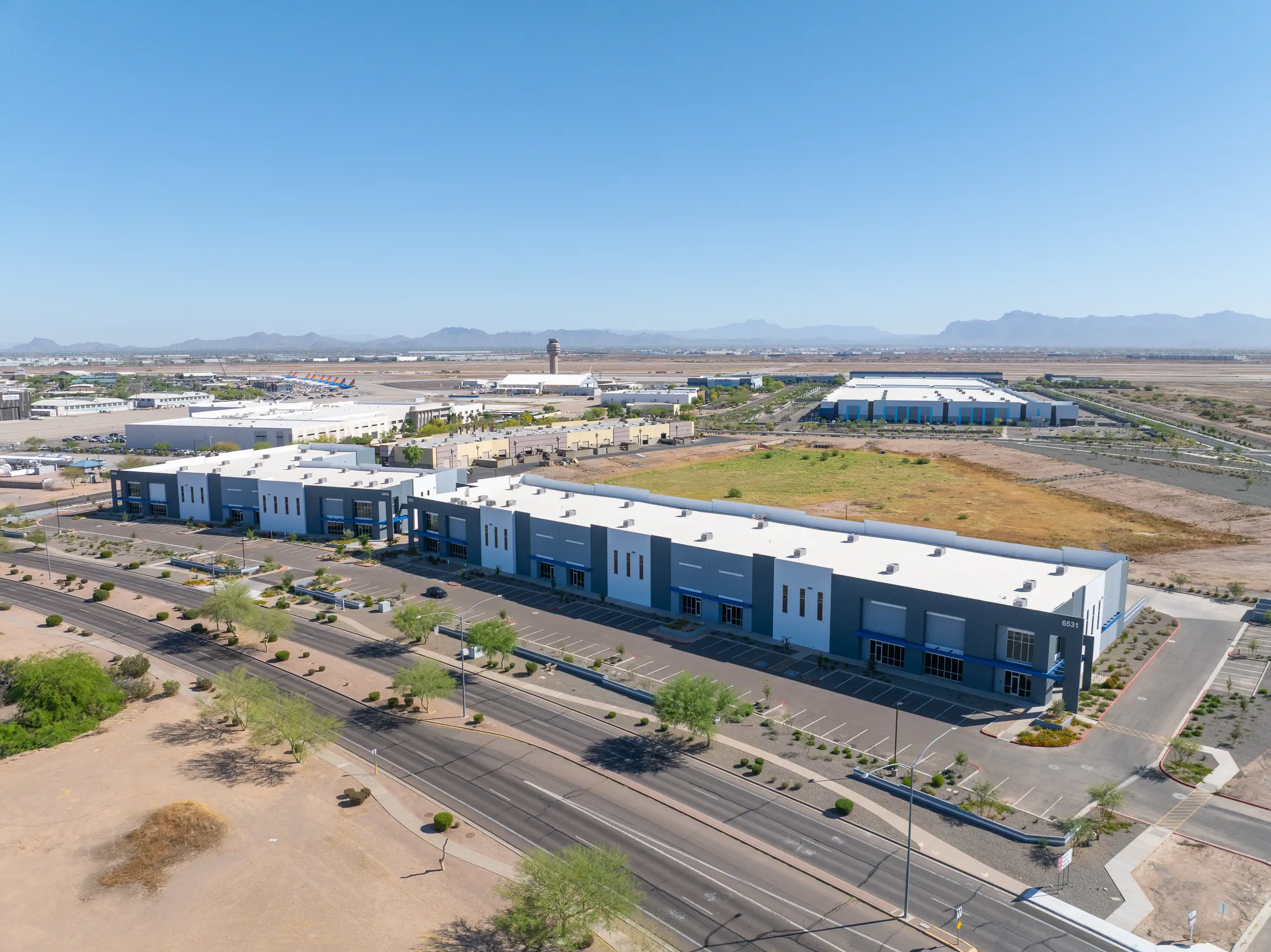 Aerial view of modern industrial buildings with parking lots next to an airport, runways, and parked planes, with mountains in the distance.