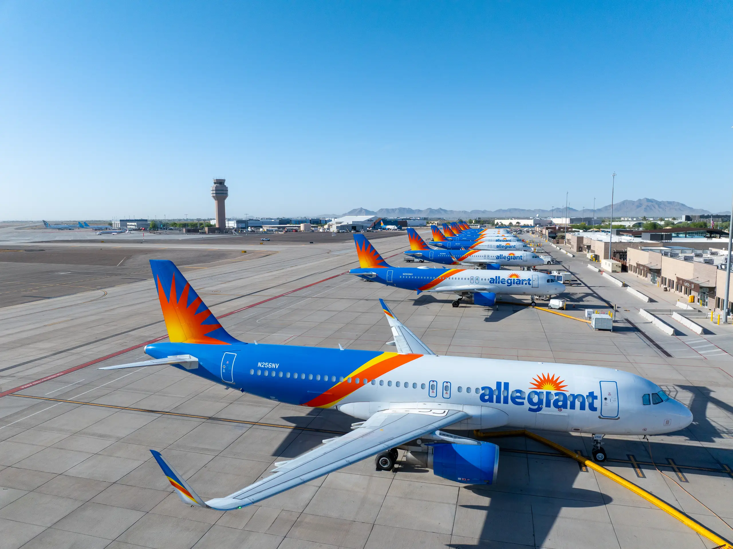 A line of blue, white, and orange Allegiant Air planes parked on an airport tarmac with a control tower and mountains in the background.