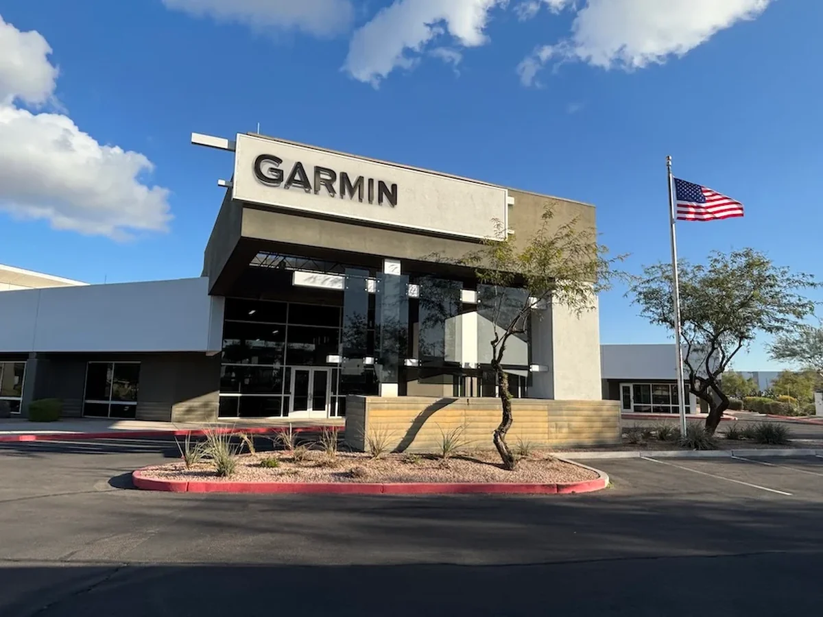 Exterior view of a modern Garmin building with large glass windows and a prominent sign, an American flag on a pole, and a landscaped foreground under a blue sky.