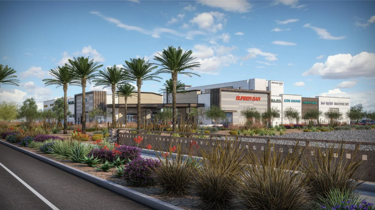 A modern commercial development with multiple storefronts, palm trees, and desert landscaping under a blue sky.