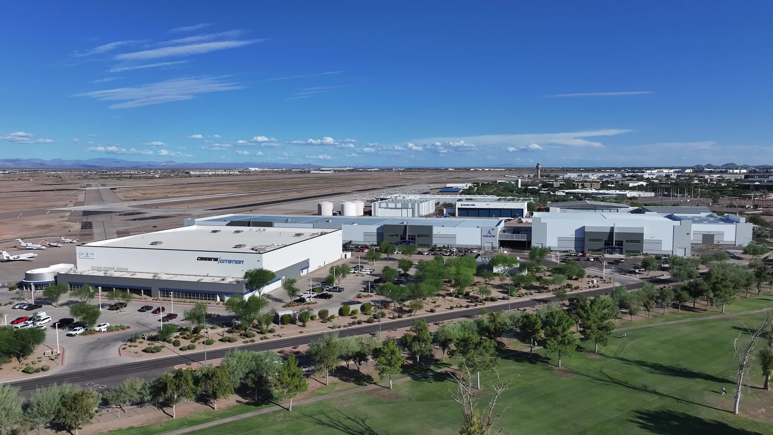 Aerial view of an airport with runways, parked jets, and large industrial buildings featuring Cessna Citation, Embraer, and Galactic logos, bordered by a green golf course and distant mountains under a blue sky.
