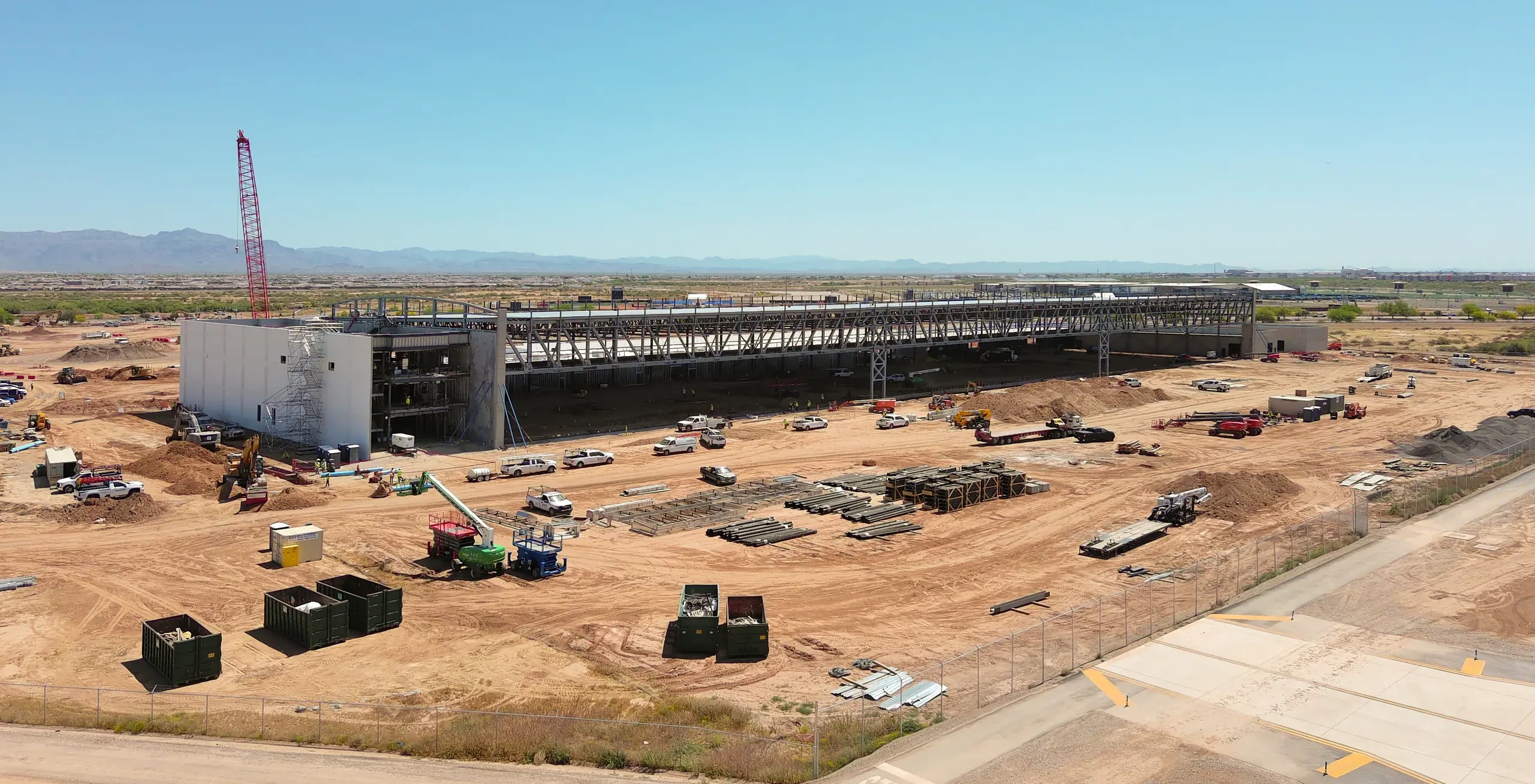 Aerial view of a large construction site with a partially built steel and concrete building, red tower crane, and various construction vehicles on dirt ground under a clear sky.
