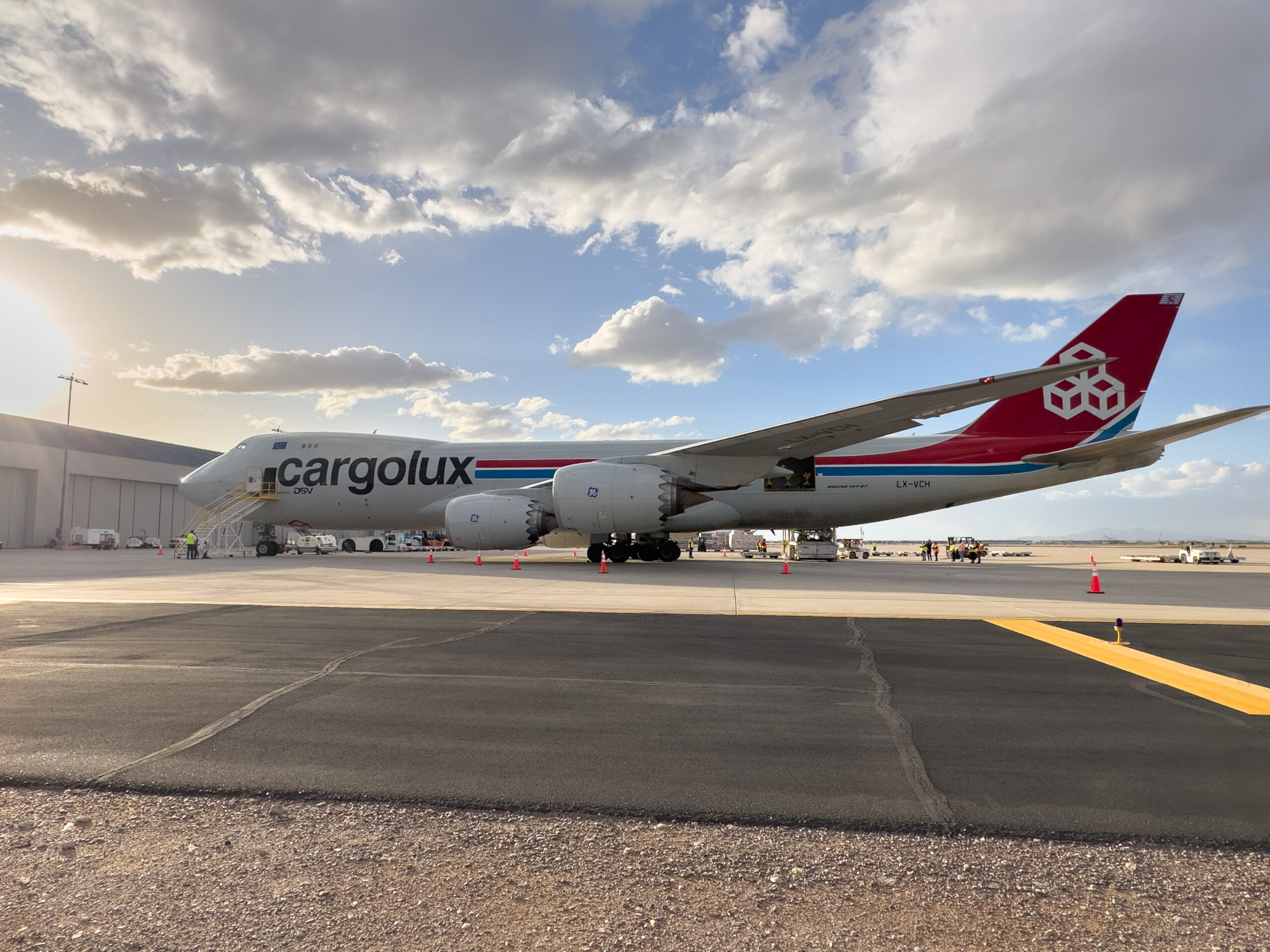 A large gray Cargolux Boeing 747-8F cargo plane with red and blue markings parked on an airport tarmac with ground crew and equipment.