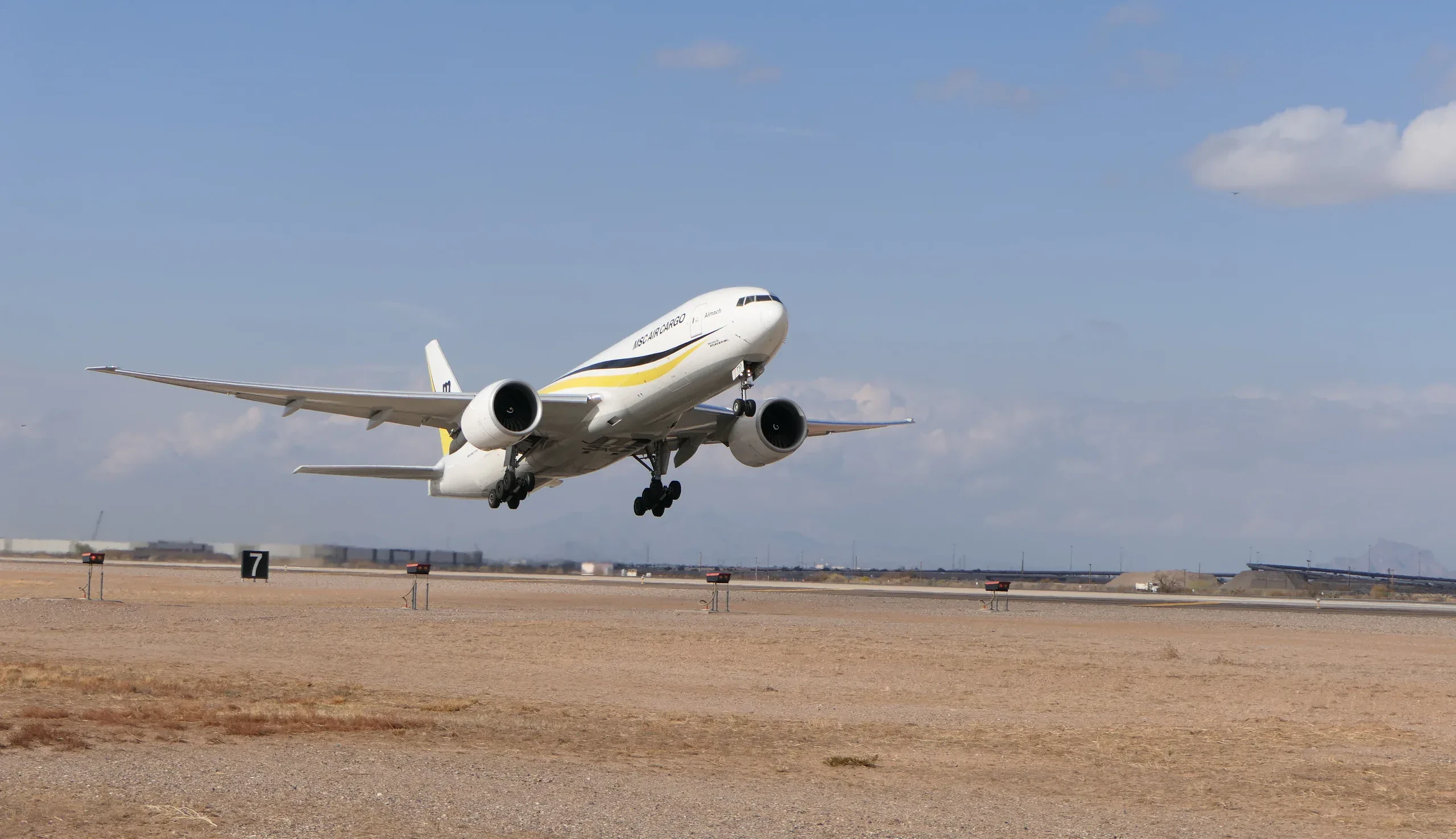 A white cargo plane with yellow and black stripes, labeled 'MSC AIR CARGO' and 'Almach', takes off from an arid airport runway under a clear blue sky.