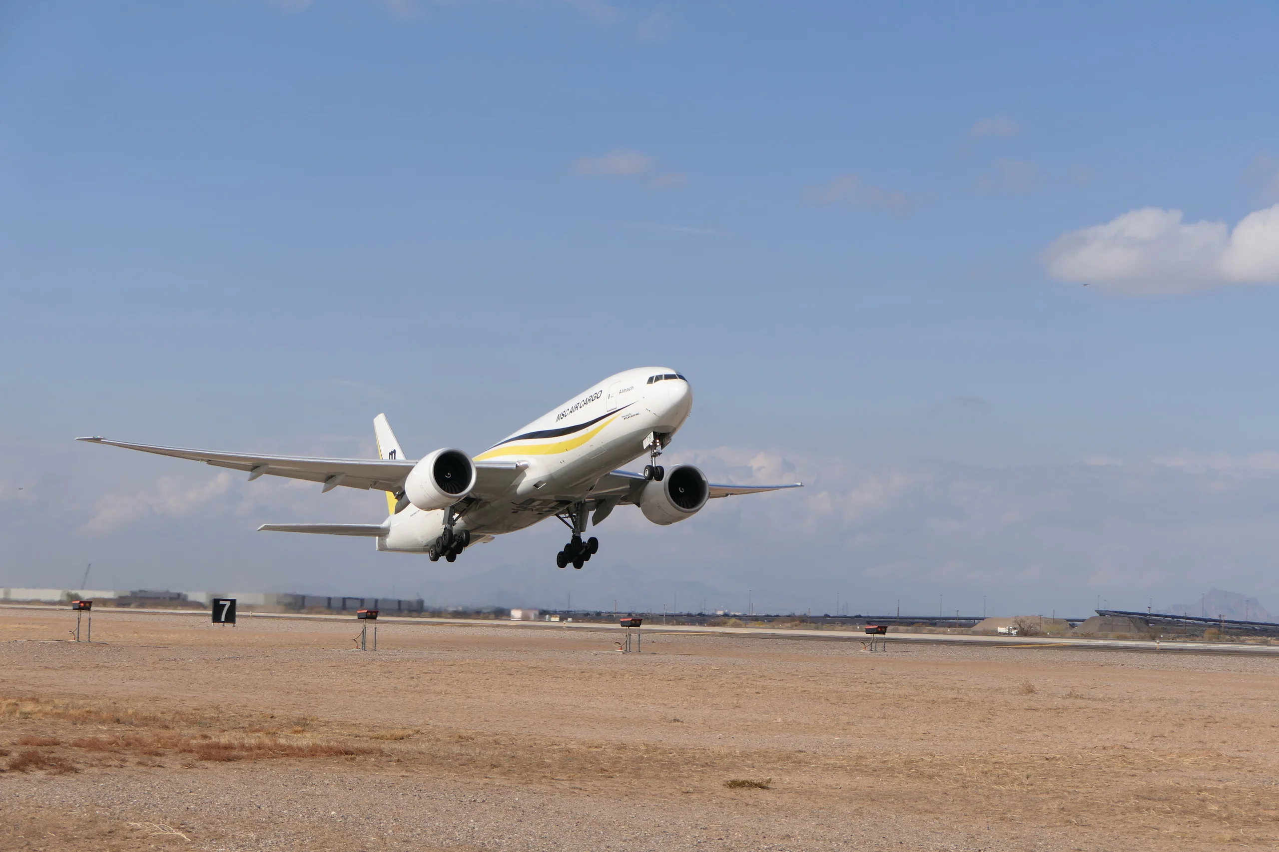 A white MSC Air Cargo Boeing 777 freighter with yellow and black stripes takes off from a runway, landing gear extended, against a blue sky.