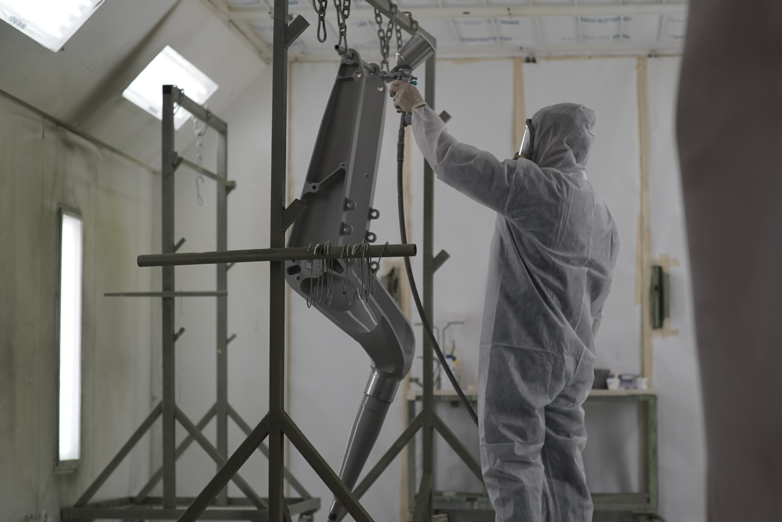 A person in a white protective suit and respirator mask spray painting a large grey metal part suspended from chains.
