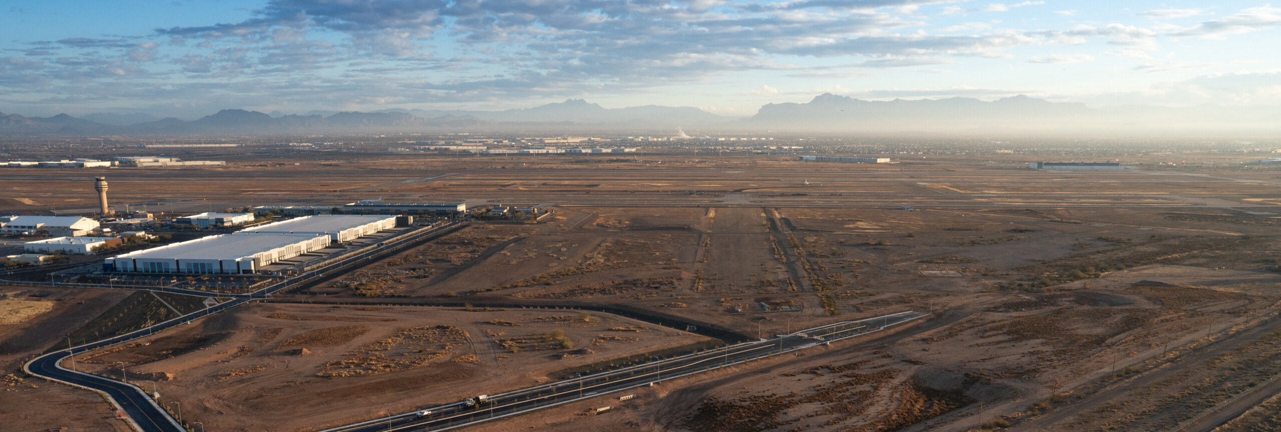 Aerial view of a desert airport and industrial complex with large buildings, runways, a control tower, and distant mountains under a cloudy sky.