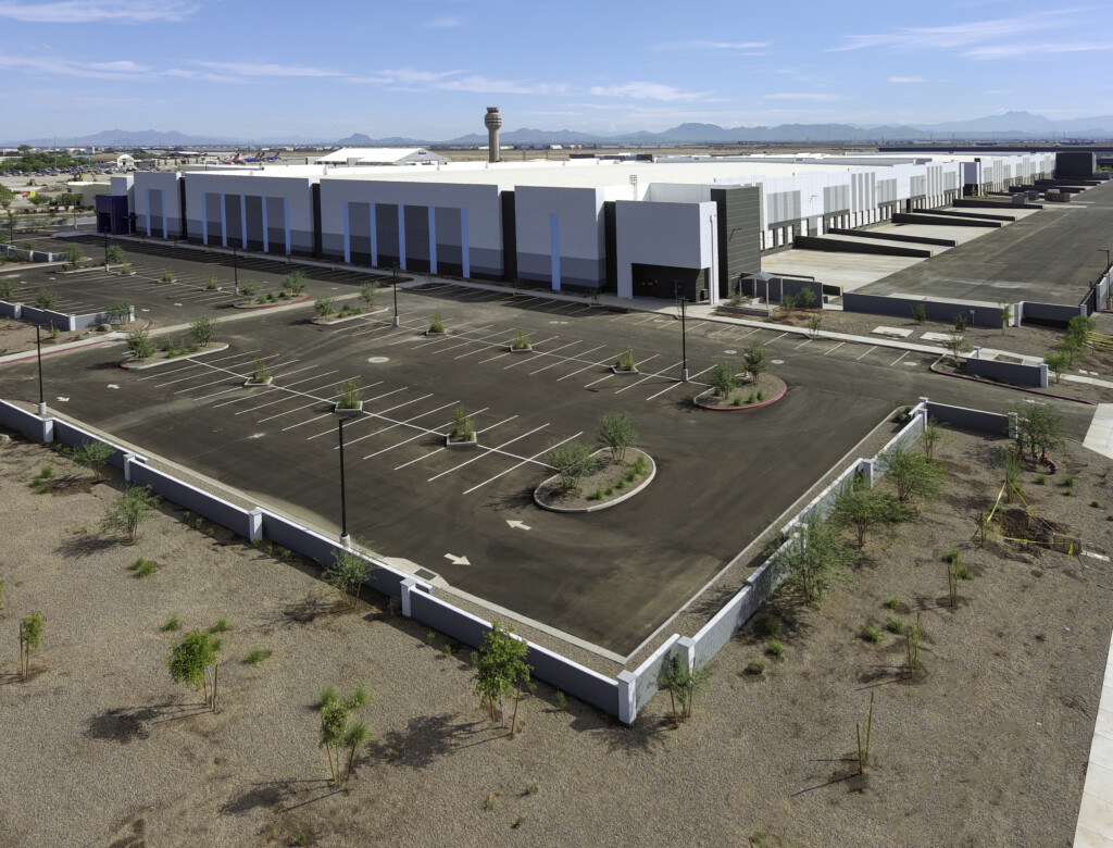 Aerial view of a large, modern industrial building with an empty parking lot, adjacent to an airport with planes and a control tower, under a blue sky.