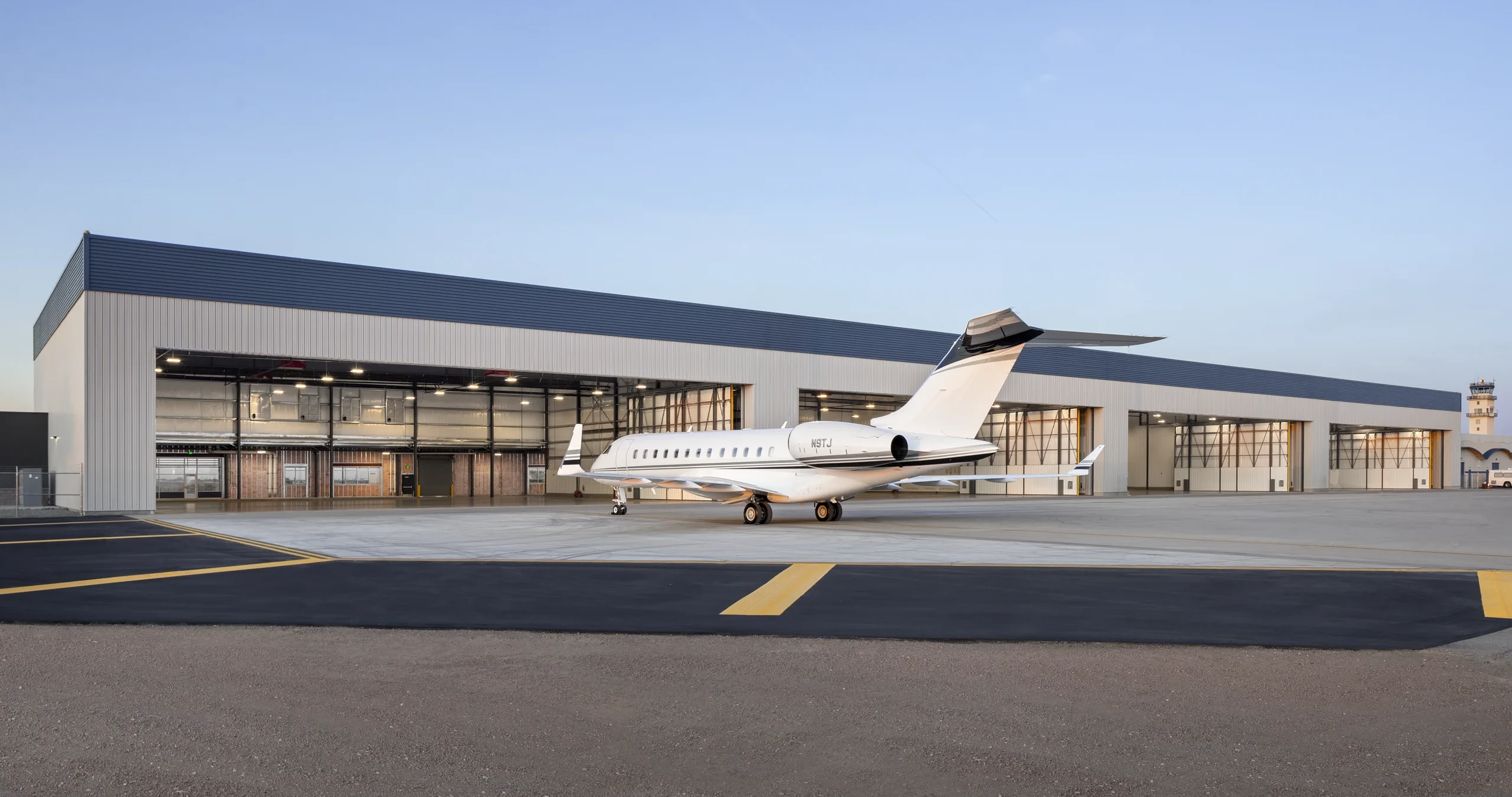 A white private jet with black and gray accents parked on a concrete apron in front of a long, modern hangar building with a dark blue roof. An air traffic control tower is visible in the distance.