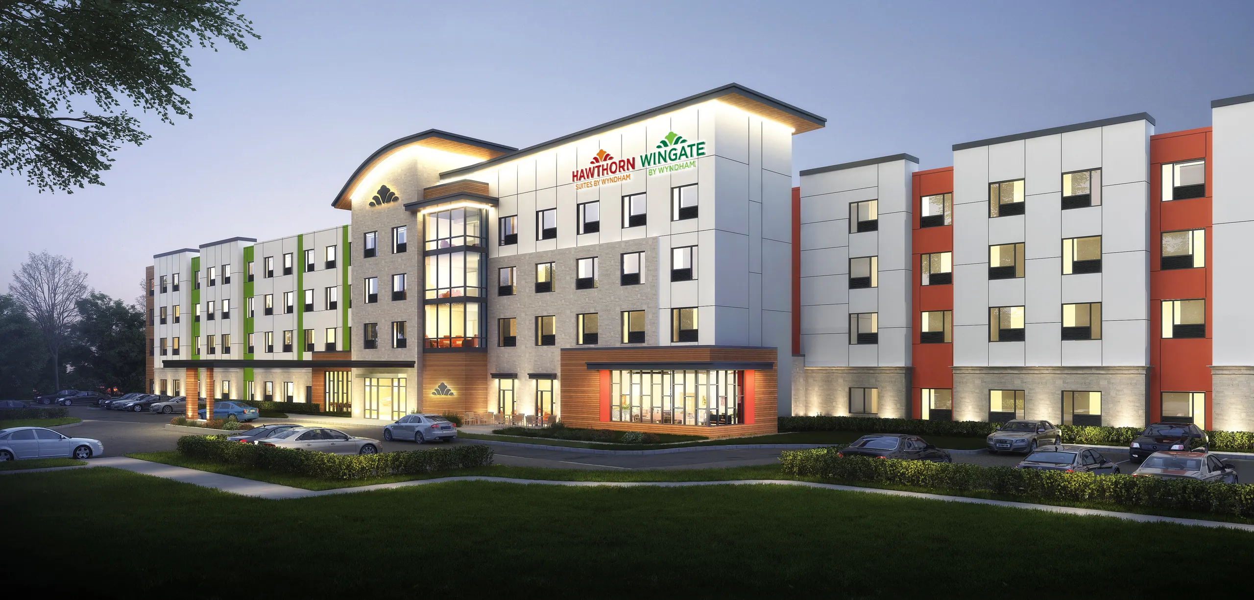 Exterior view of a modern Hawthorn Wingate Suites by Wyndham hotel at dusk, featuring white, gray, green, and terracotta red facades, illuminated windows, and a parking lot with cars.