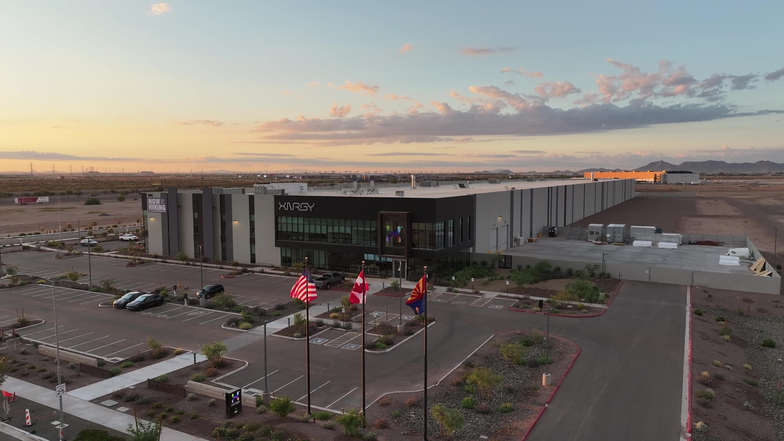 Aerial view of the XNRGY industrial building with flags, a parking lot, and desert landscaping under a colorful sunset sky.